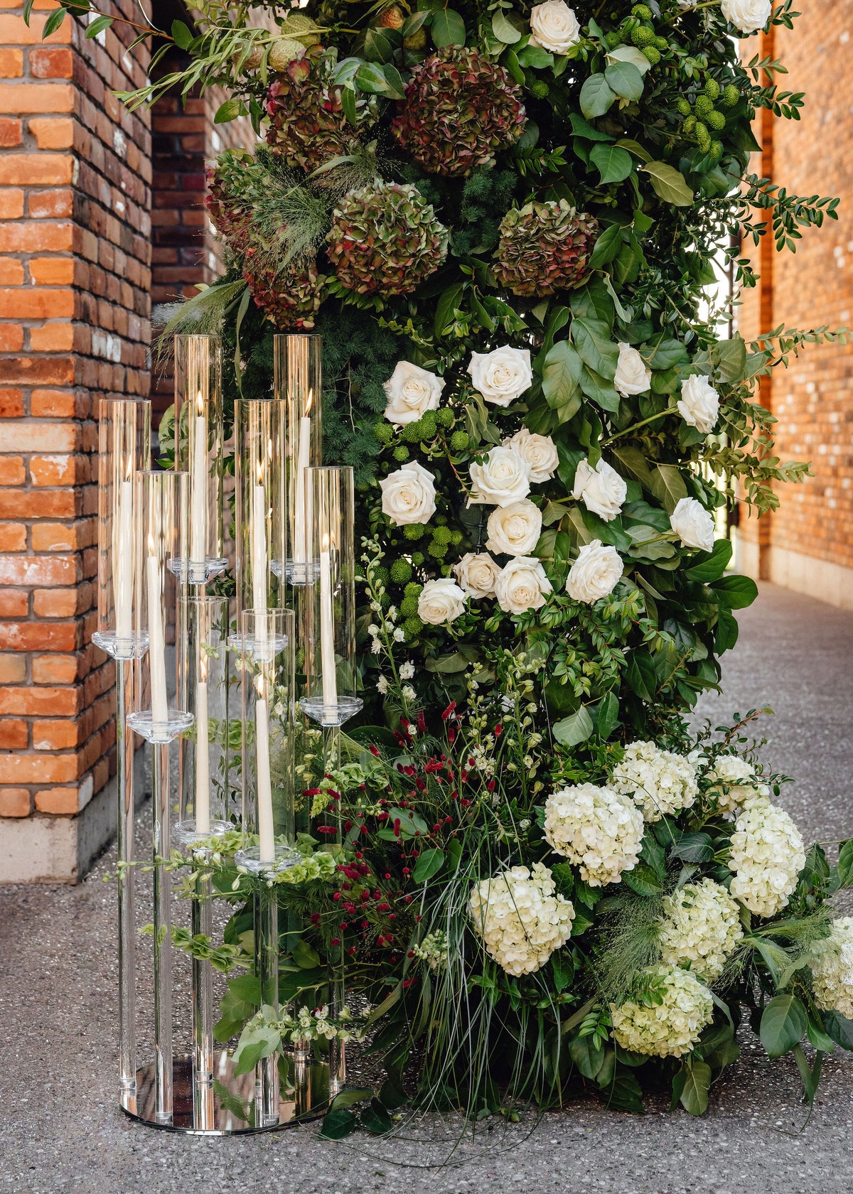 A large floral wall arrangement with white roses, white hydrangeas, green foliage, and dried greenery, paired with a tall mirrored glass candle holder, set outdoors against a brick wall.