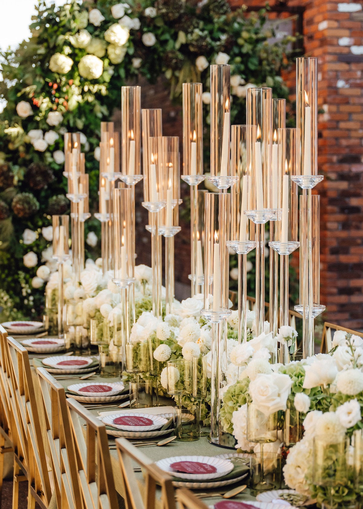 A long table set for guests with tall clear glass candlestick holders, white rose centerpieces, white plates, and a rustic brick wall and floral backdrop.