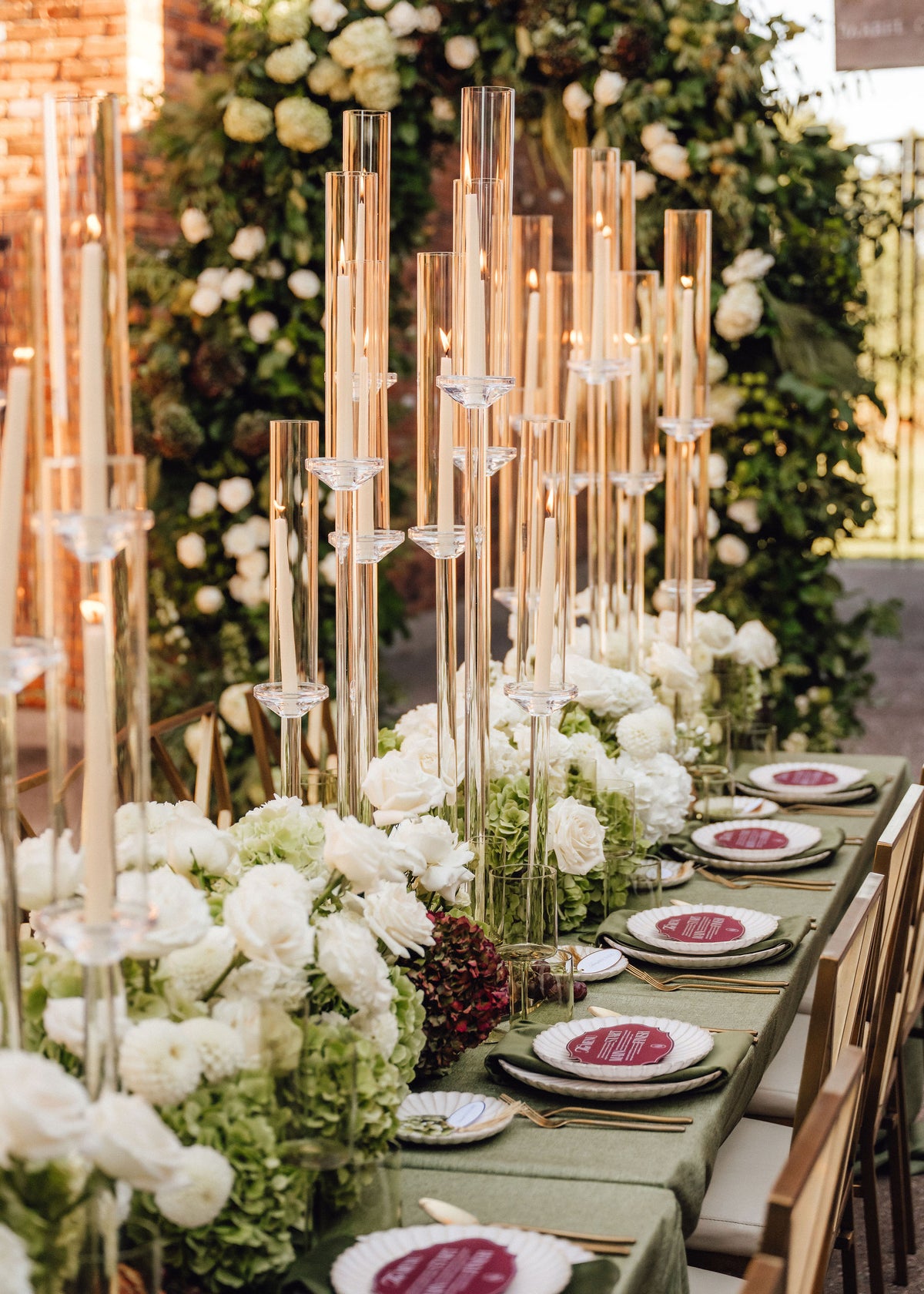 A long event table decorated with tall clear glass candlestick holders, white rose and hydrangea floral arrangements, white plates, and a green tablecloth in an outdoor venue setting.