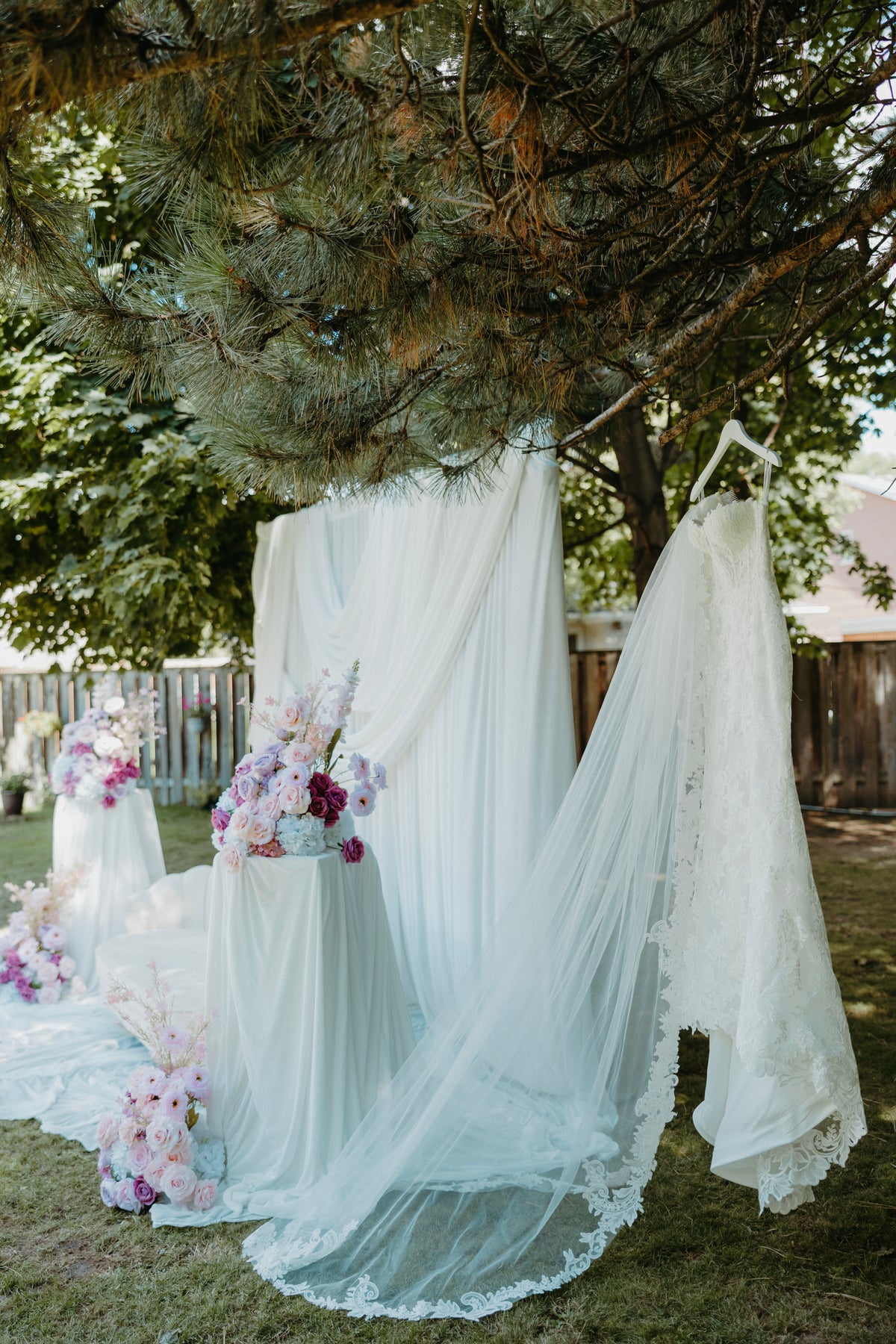 A white wedding dress with a long sheer veil and lace-trimmed hem is displayed outdoors under a tree, alongside floral arrangements and a matching veil setup on a lawn.