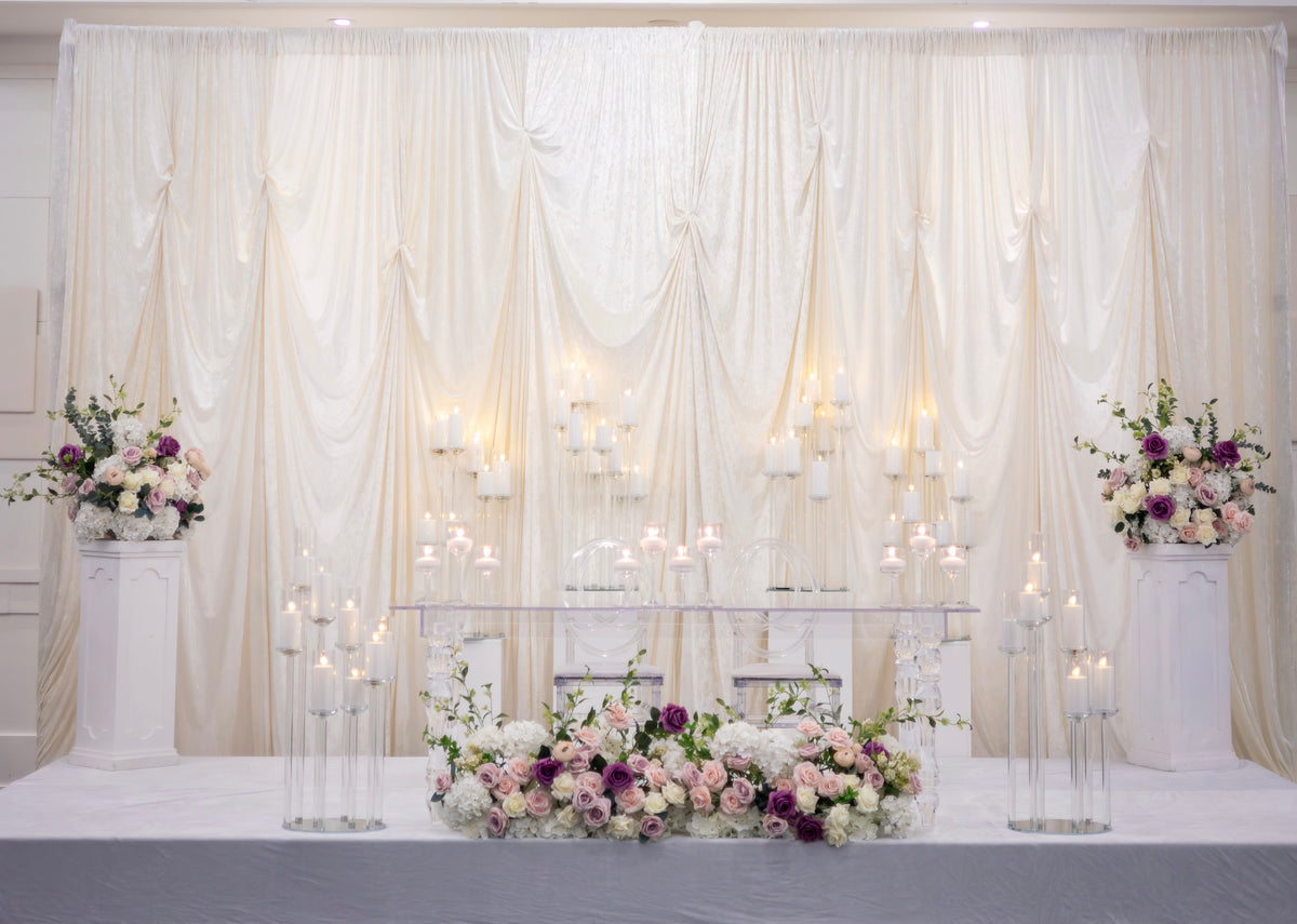 A decorated event backdrop with draped cream fabric, multiple white candelabras with lit candles, a long floral arrangement in the foreground, and matching floral pedestals on both sides.