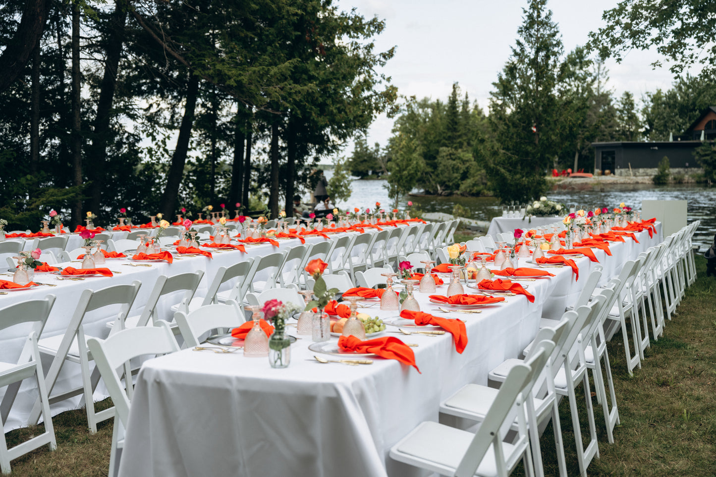 Outdoor wedding setup with white chairs and tables draped in white tablecloths with red napkins, surrounded by trees and a body of water.
