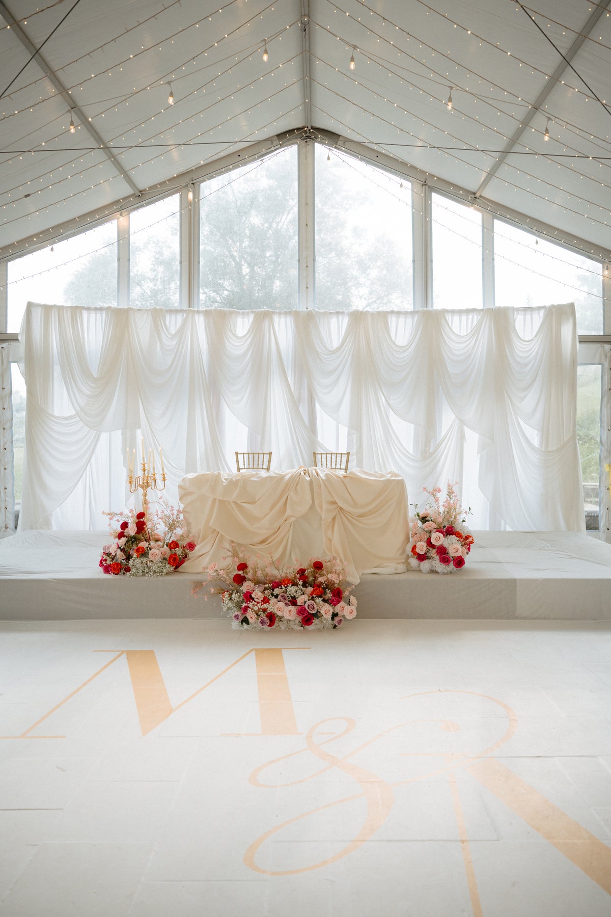 A wedding reception setup inside a white marquee tent, featuring a draped sweetheart table with gold chairs, red and white floral arrangements, and a white draped backdrop. Gold monogram letters are printed on the floor.