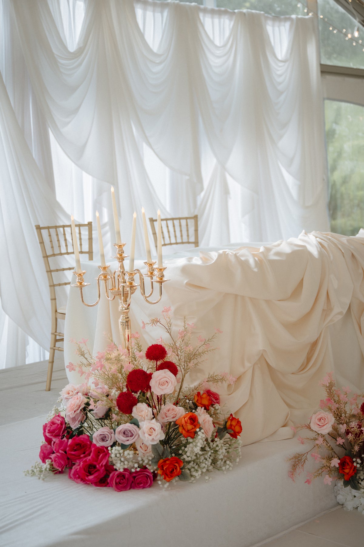 A styled bridal or event decor scene with a draped cream satin bed or sofa, white sheer curtains, gold chairs, a gold candelabra, and floral arrangements in pink, red, orange, and white in the foreground.