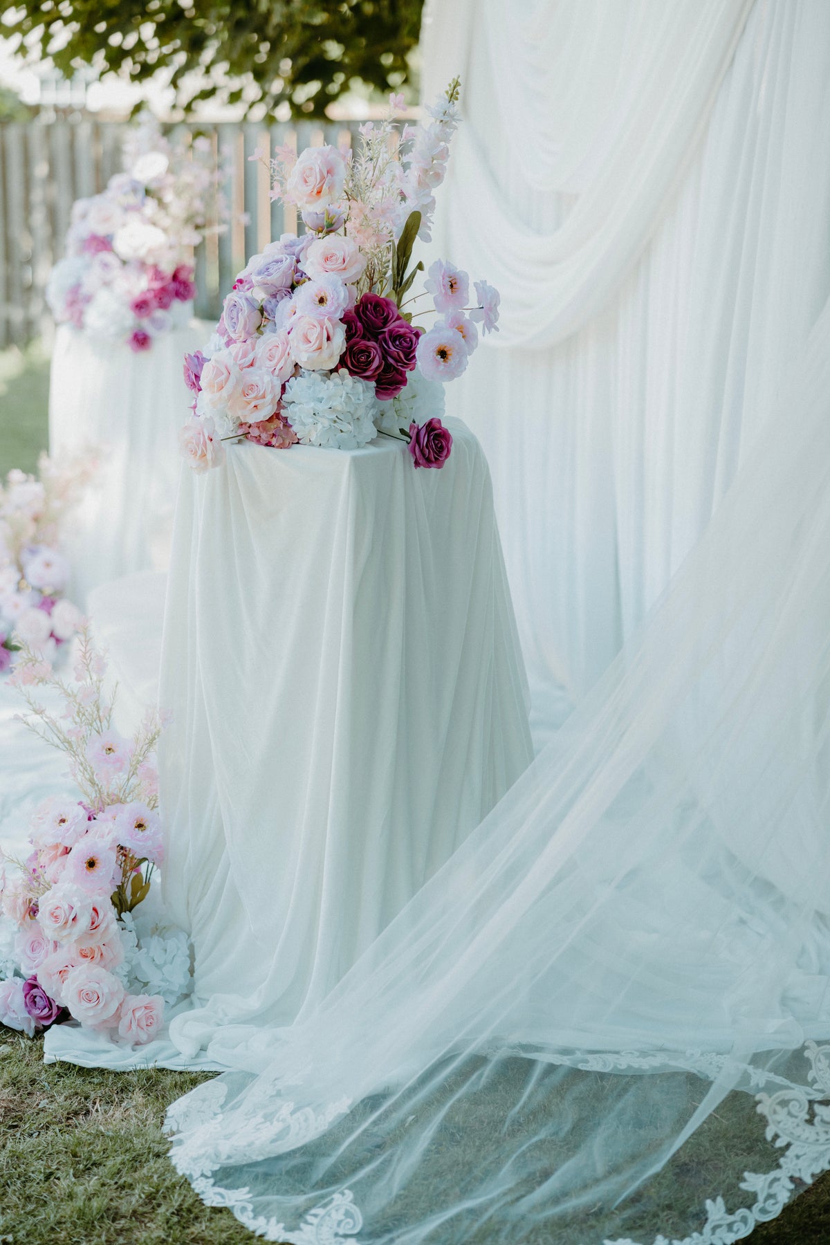 A draped white fabric setup outdoors with a large veil or tulle fabric spread across the ground and on a pedestal, accented by pastel and deep pink floral arrangements.