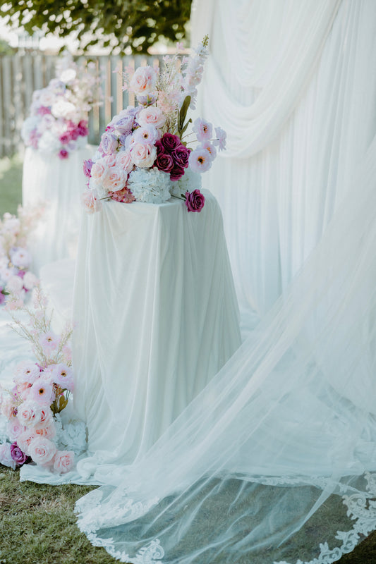 A draped white fabric setup outdoors with a large veil or tulle fabric spread across the ground and on a pedestal, accented by pastel and deep pink floral arrangements.