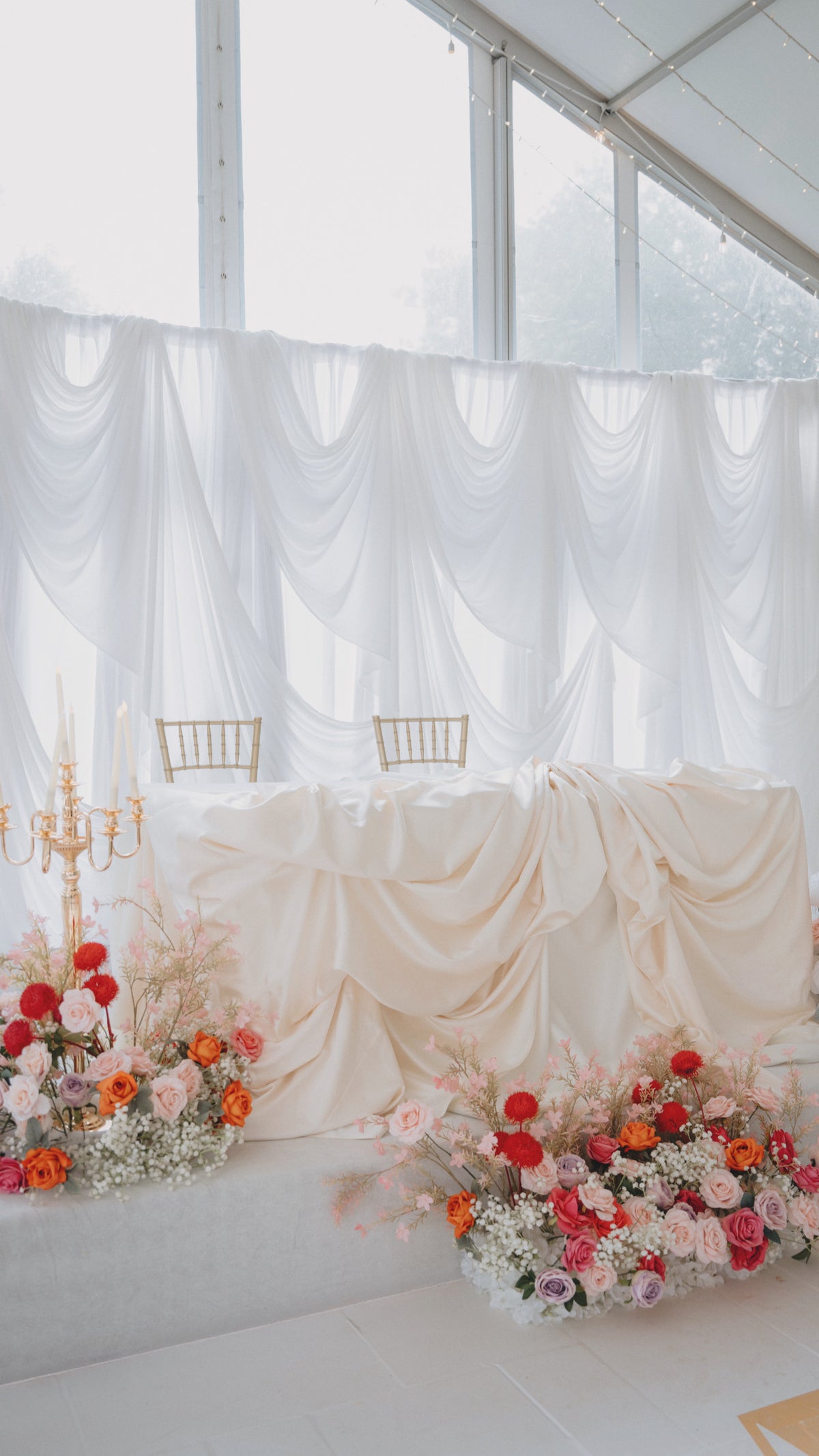 A styled wedding reception table setup with a draped cream tablecloth, gold chairs, and floral arrangements in pink, red, and orange in front of white draped fabric and large windows.