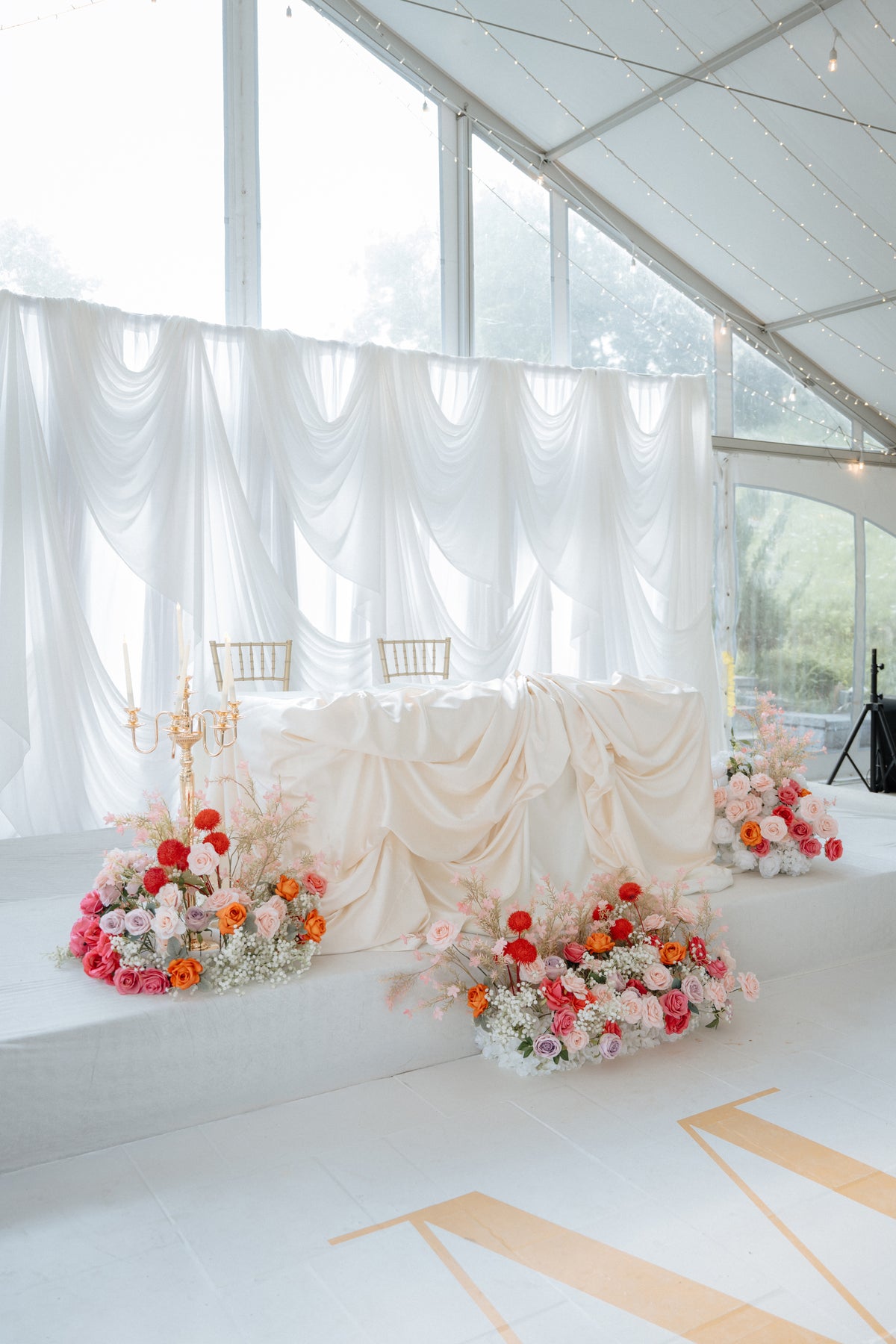 A decorated event stage inside a white tent with draped fabric, white draped table covering, gold chairs, and multiple floral arrangements in pink, orange, and white on the floor.