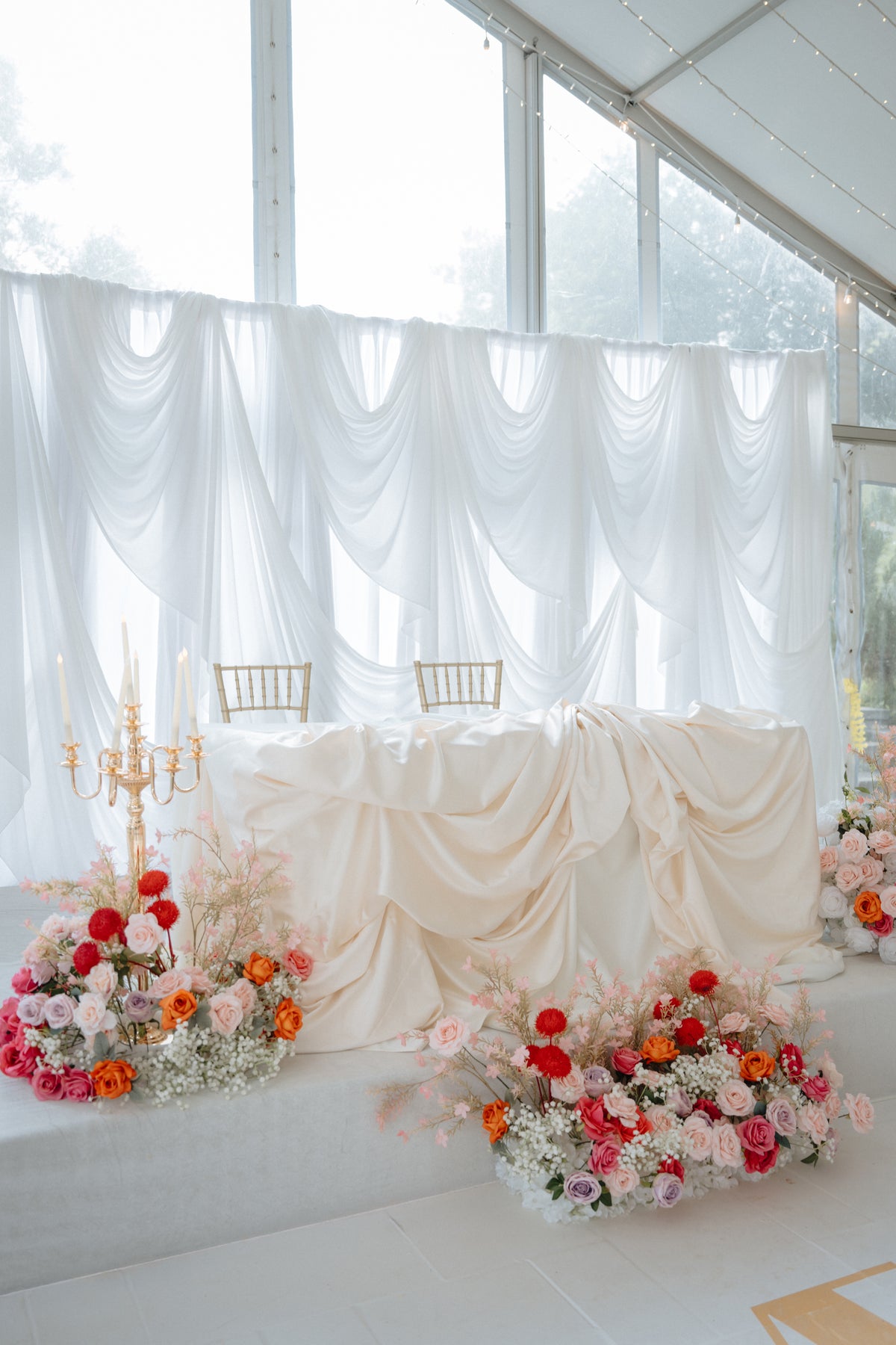 A decorated event table draped in cream fabric, set with two gold chairs, white draped curtains in the background, and colorful floral arrangements in pink, red, and orange at the front.