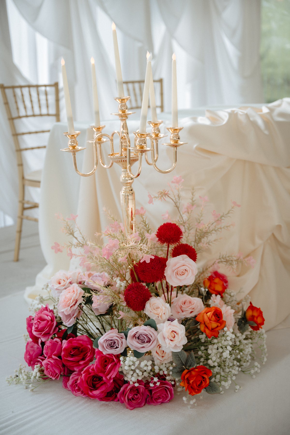 A large floral centerpiece arrangement with pink, red, peach, and lavender roses, baby's breath, and filler stems in front of a draped cream tablecloth, with a gold candelabra and chairs in the softly lit background.