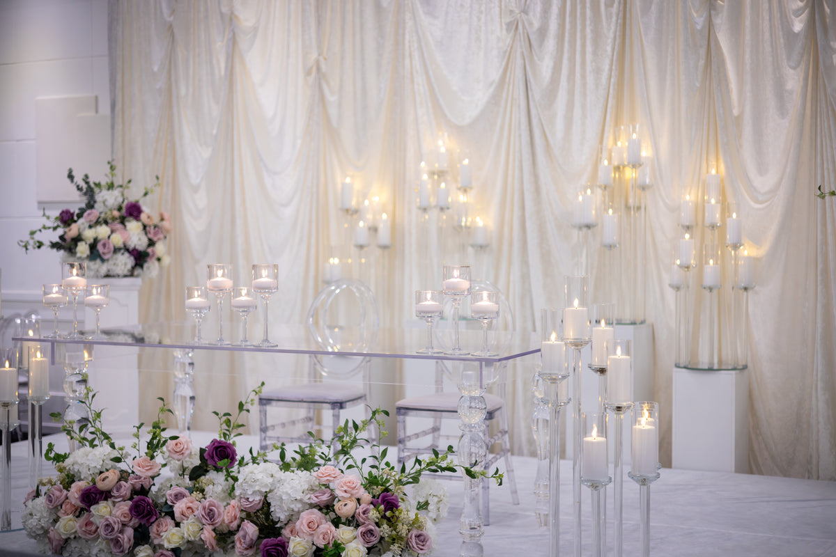 A wedding or event backdrop setup with draped cream fabric, multiple clear acrylic candle holders with lit candles, and floral arrangements in white, pink, and purple in the foreground and on a pedestal.