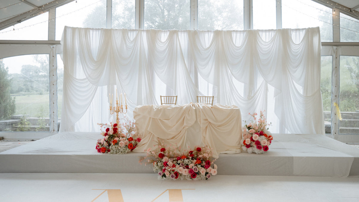 A decorated event stage inside a glass-roofed pavilion with white draped fabric backdrops, a draped table in the center, two chairs, floral arrangements in red, pink, and white, and a white floor with large gold letters.