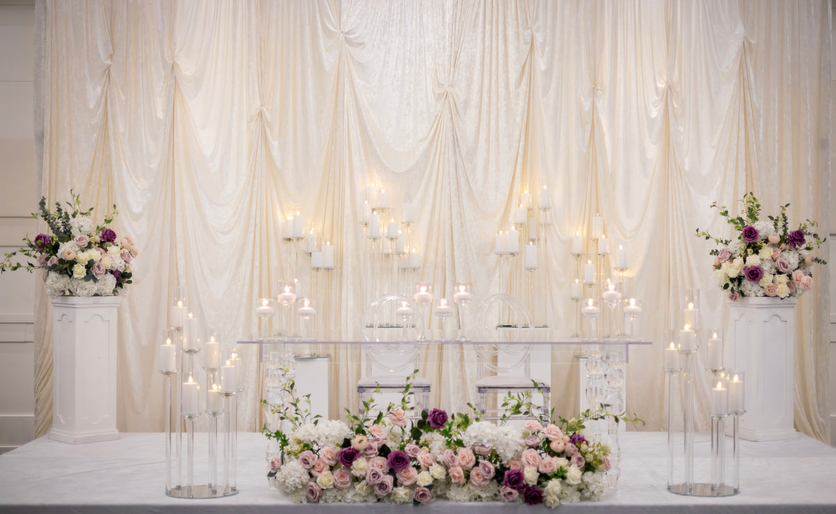 A wedding or event backdrop setup featuring draped cream fabric curtains, clear glass candelabras with lit candles, and floral arrangements in white, blush pink, and purple on pedestals.