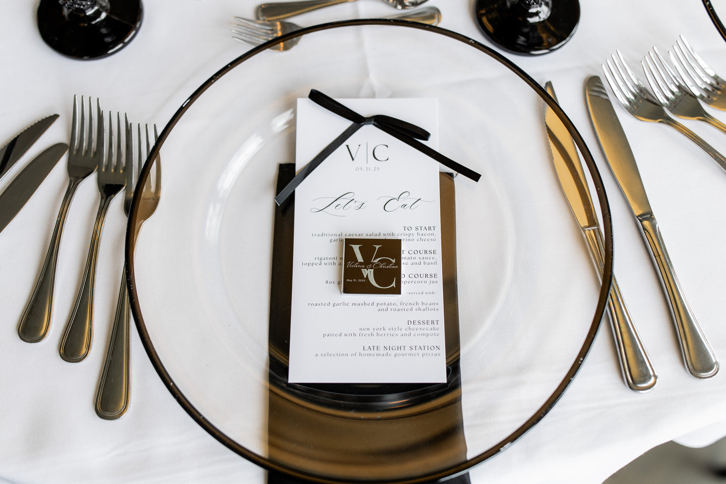 Dining table setting with a clear glass plate, black napkin, and event invitation card.