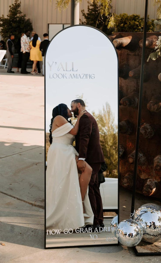 Couple in wedding attire standing next to a mirror with promotional text.