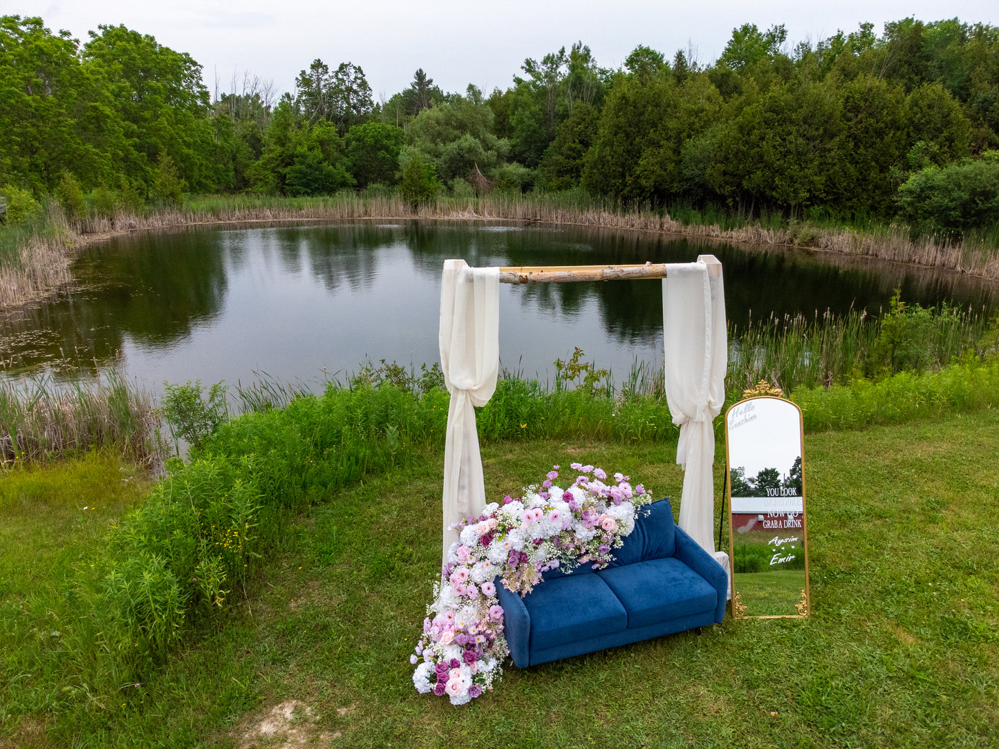 Decorative setup with a blue couch, floral arrangement, and mirror by a pond with trees in the background.