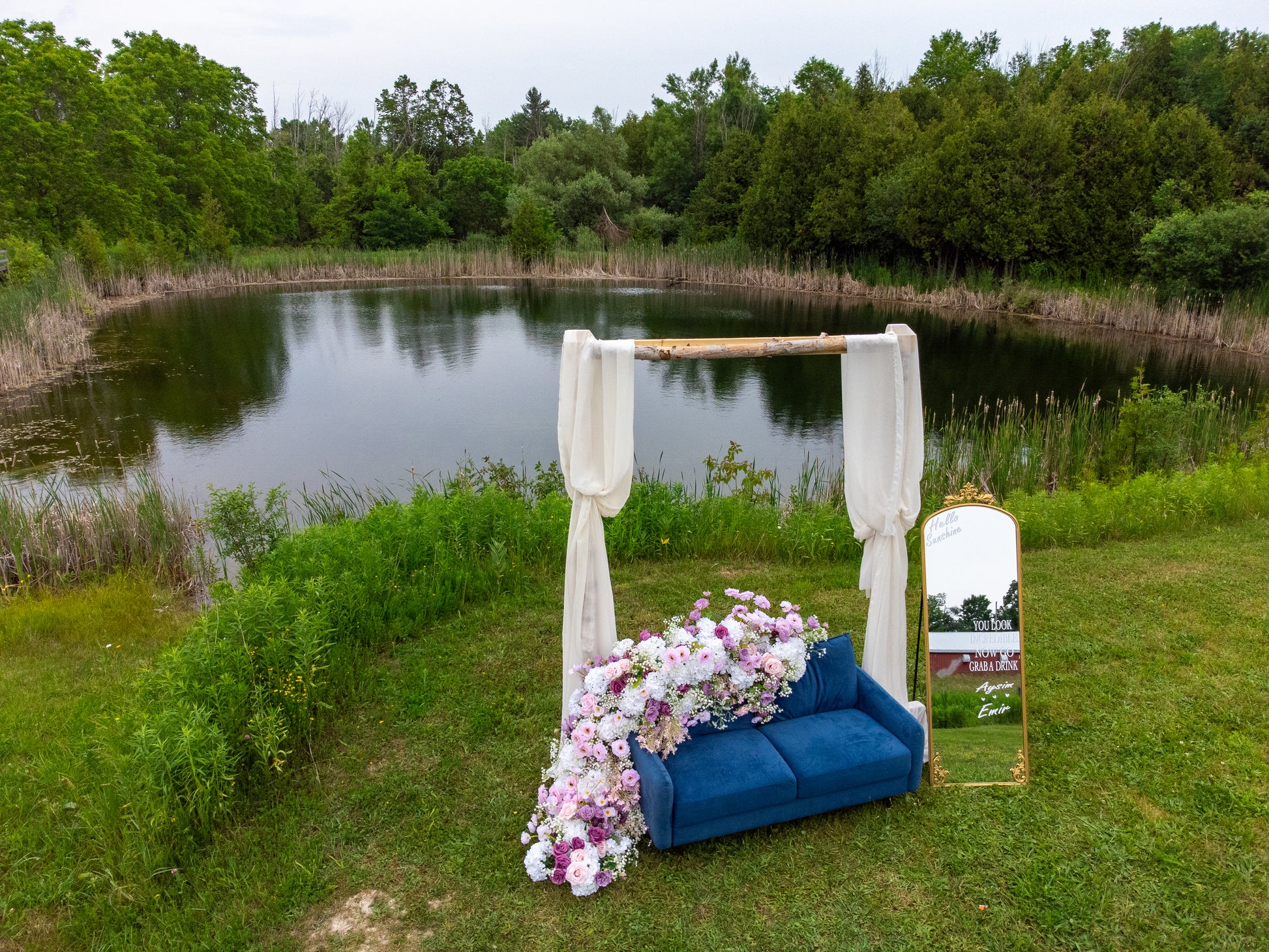 Decorative setup with a blue couch, floral arrangement, and mirror by a pond with trees in the background.