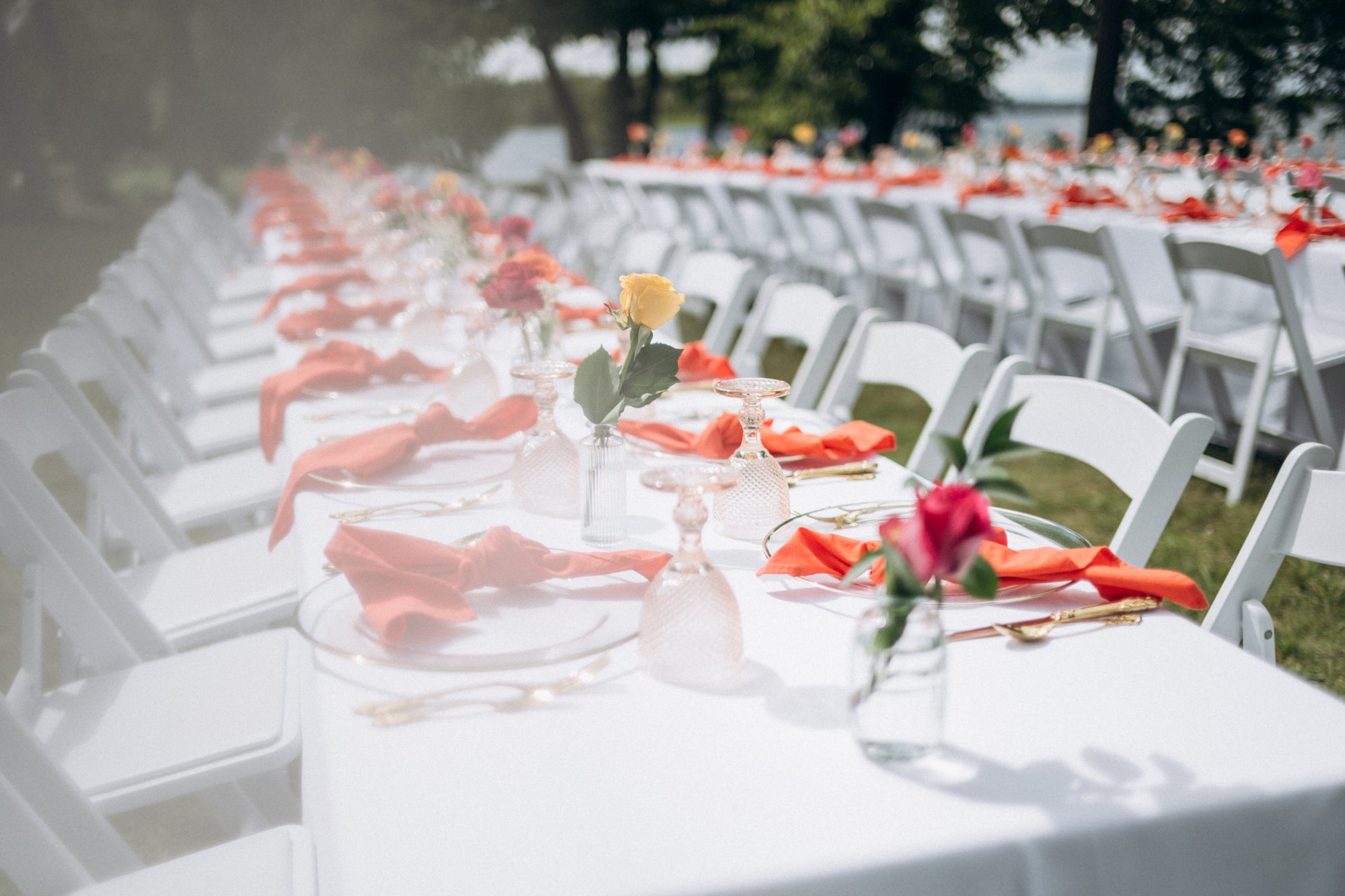 Long table set for an outdoor event with white chairs, red napkins, and floral centerpieces.