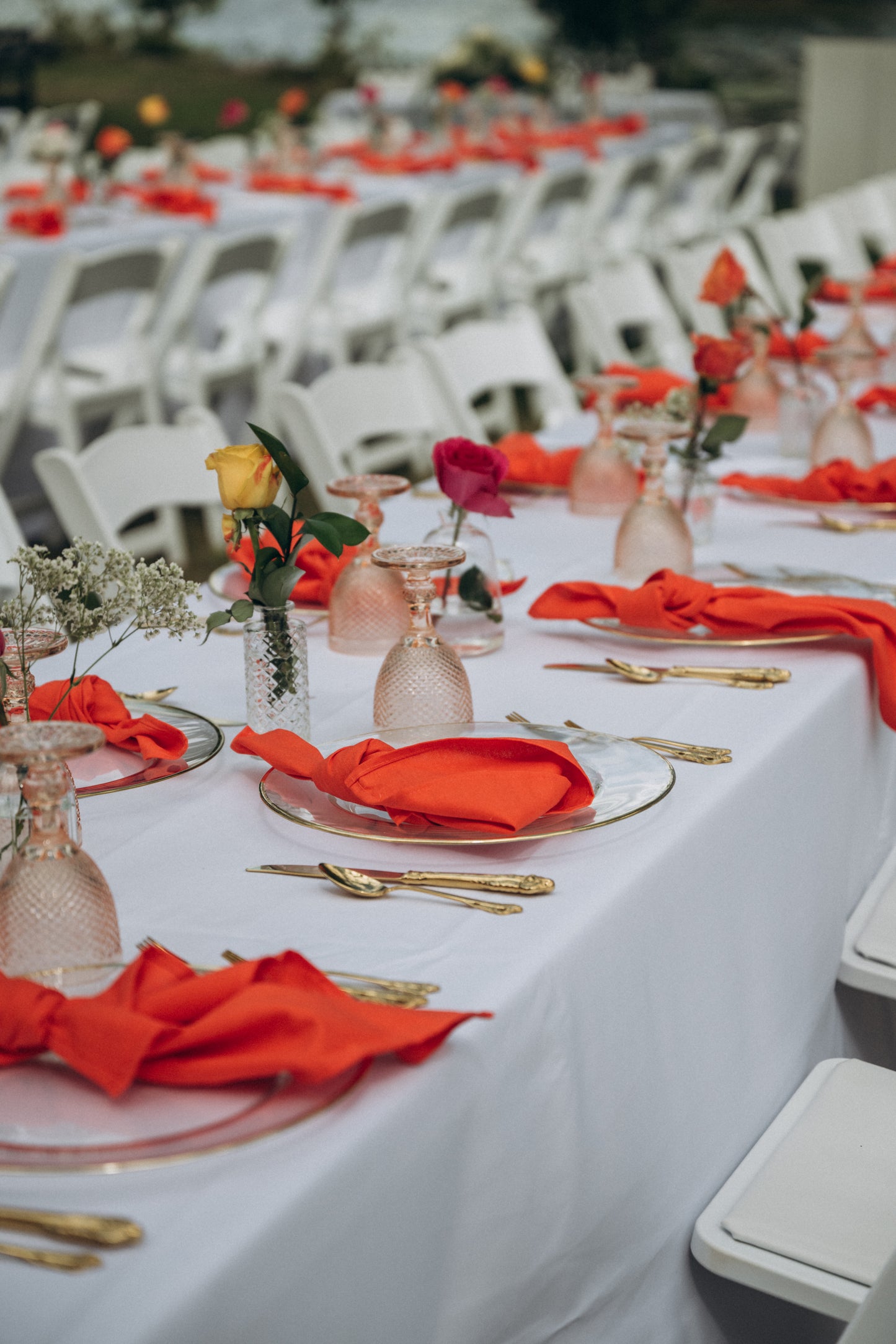 Table setting with red napkins, gold cutlery, and floral arrangements at an outdoor event.