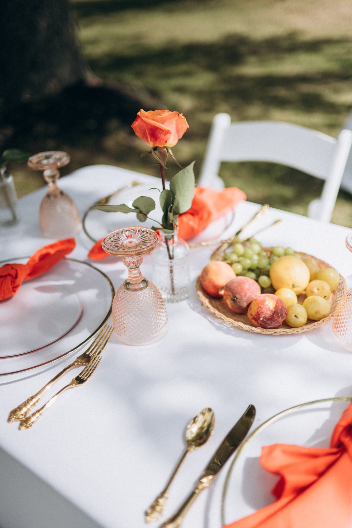 Table setting with orange napkins, fruit bowl, and rose on a white tablecloth outdoors.
