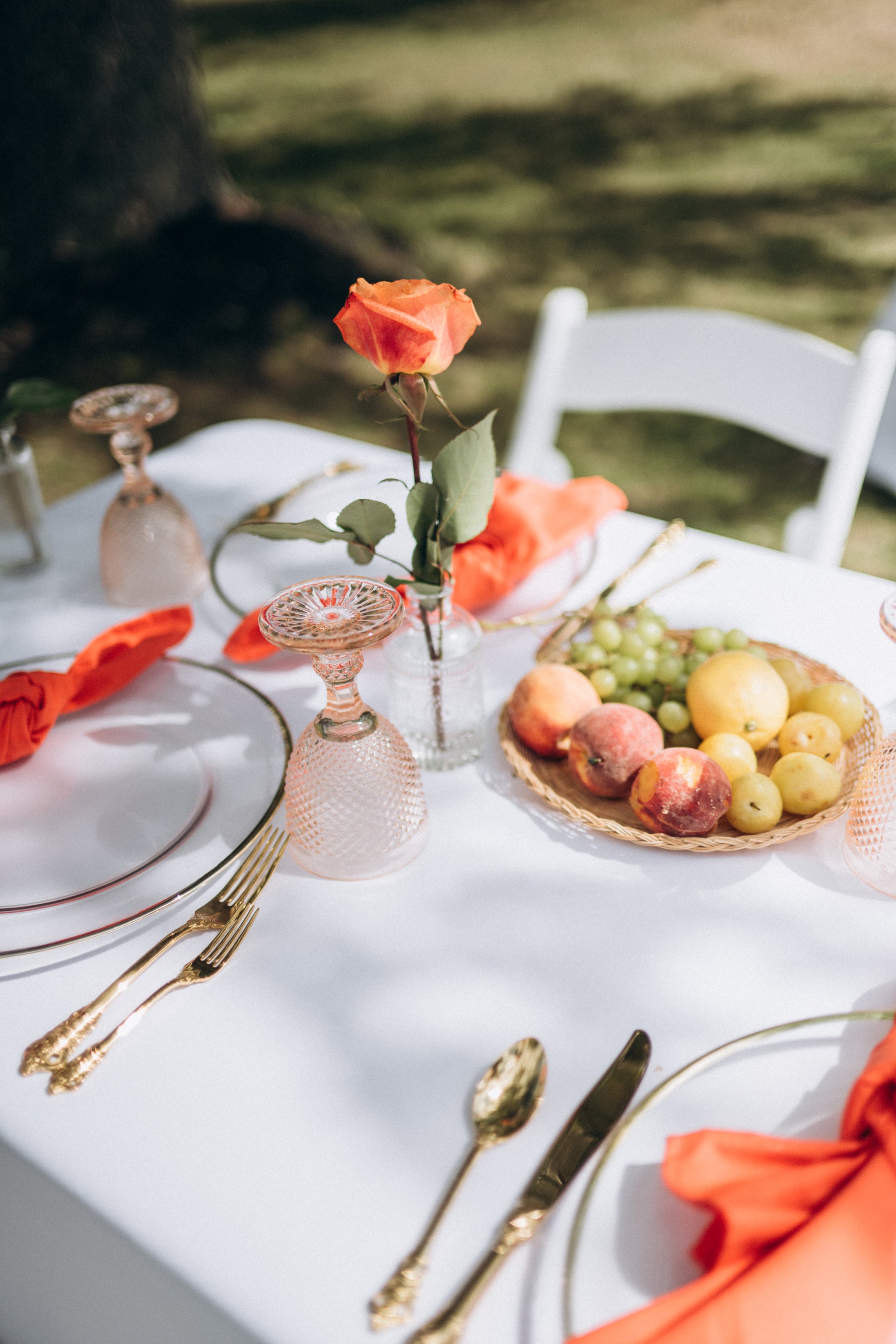 Table setting with orange napkins, fruit bowl, and rose on a white tablecloth outdoors.
