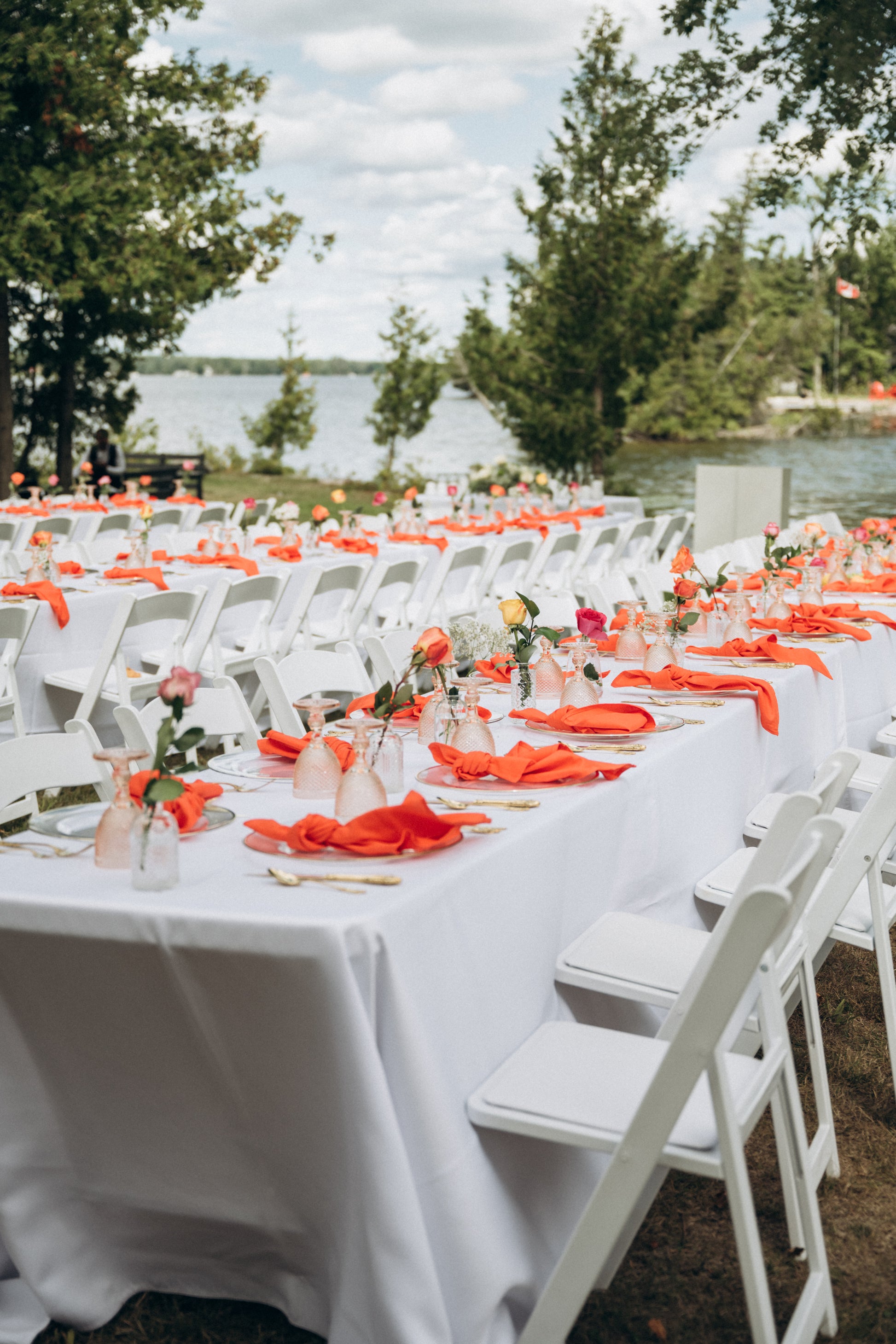 Outdoor wedding setup with white chairs, tables draped in white tablecloths, and orange napkins by a waterfront.
