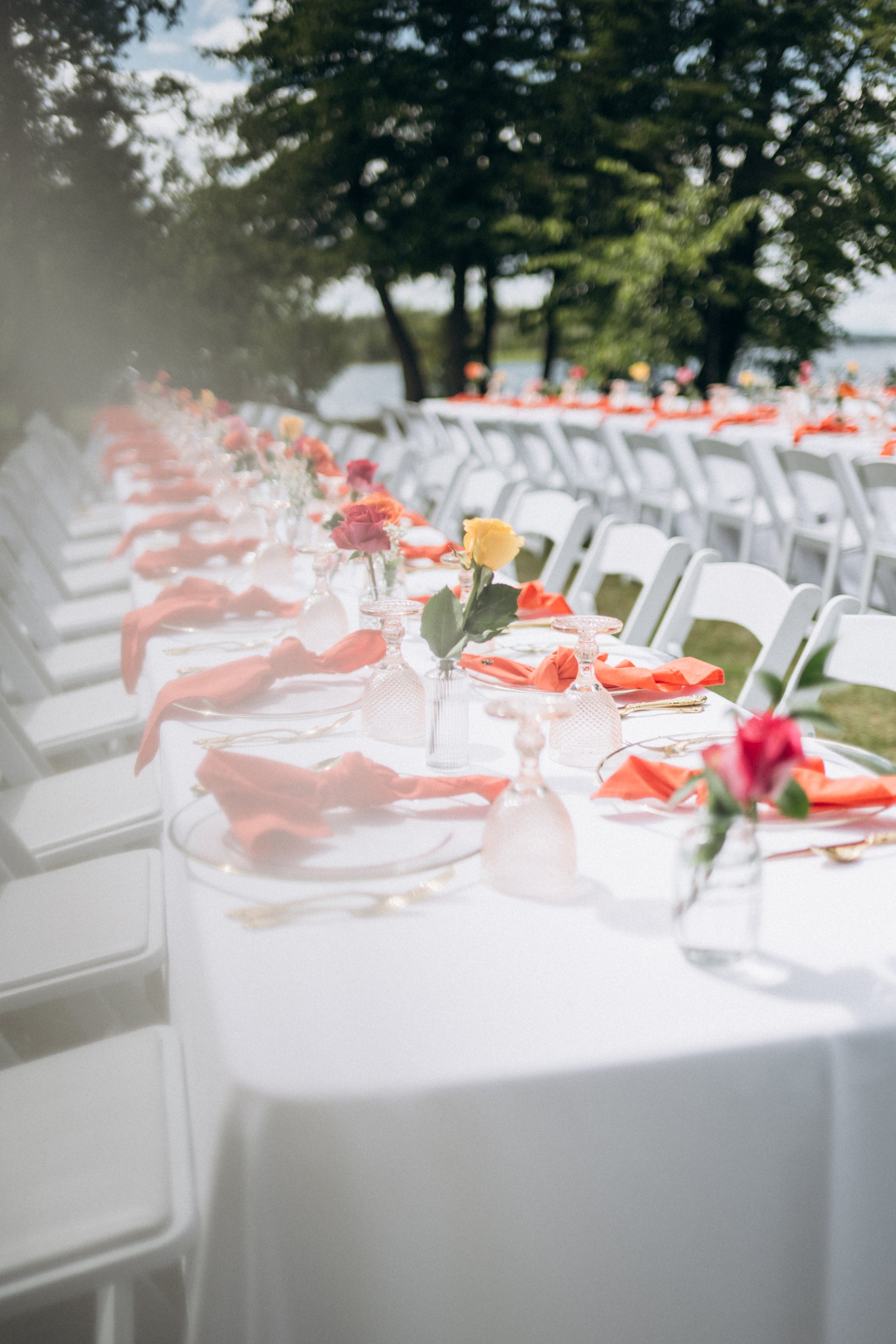 Long table set for an outdoor event with white chairs, orange napkins, and floral centerpieces.