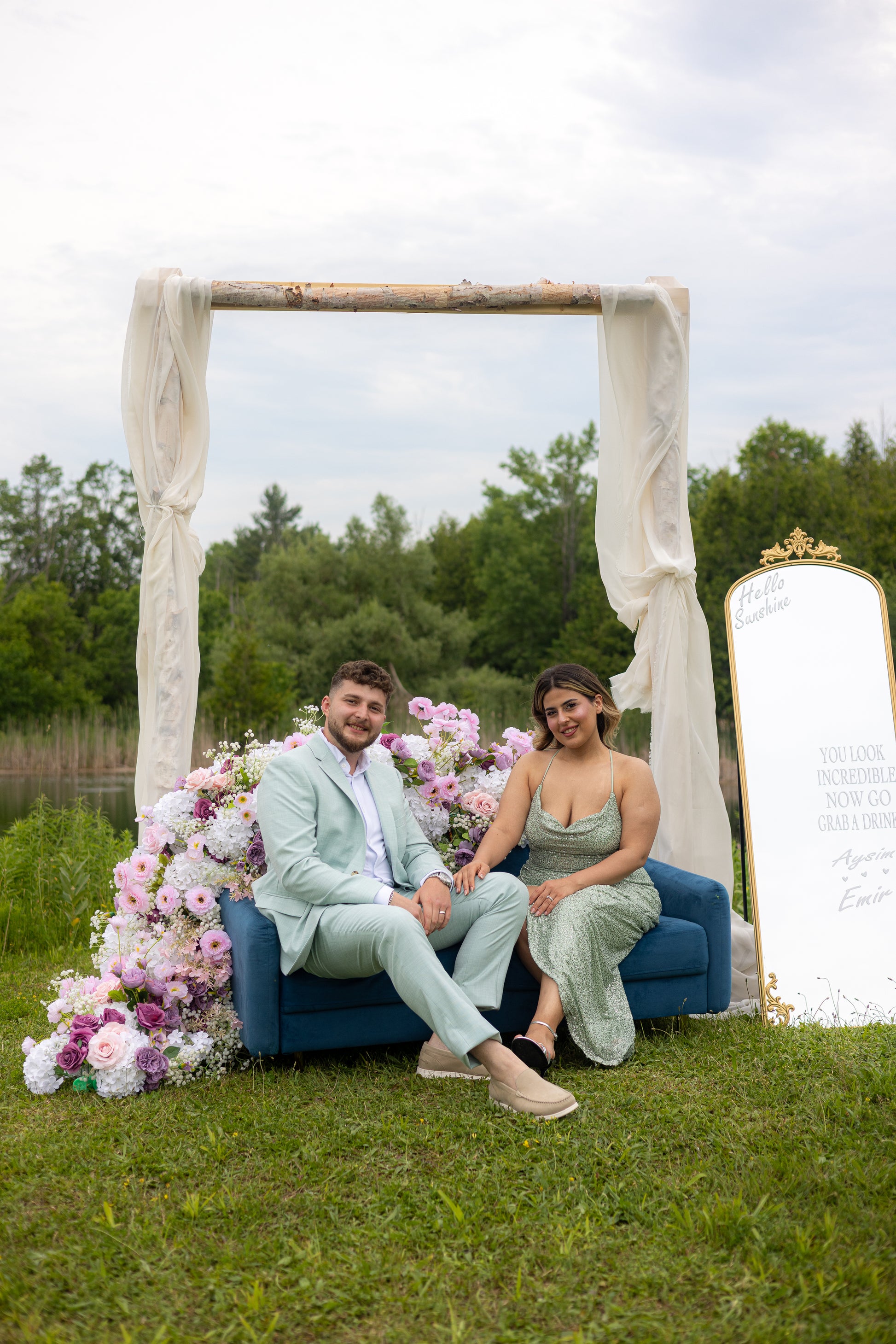 Two people sitting on a blue couch under a floral arch with a mirror in the background.