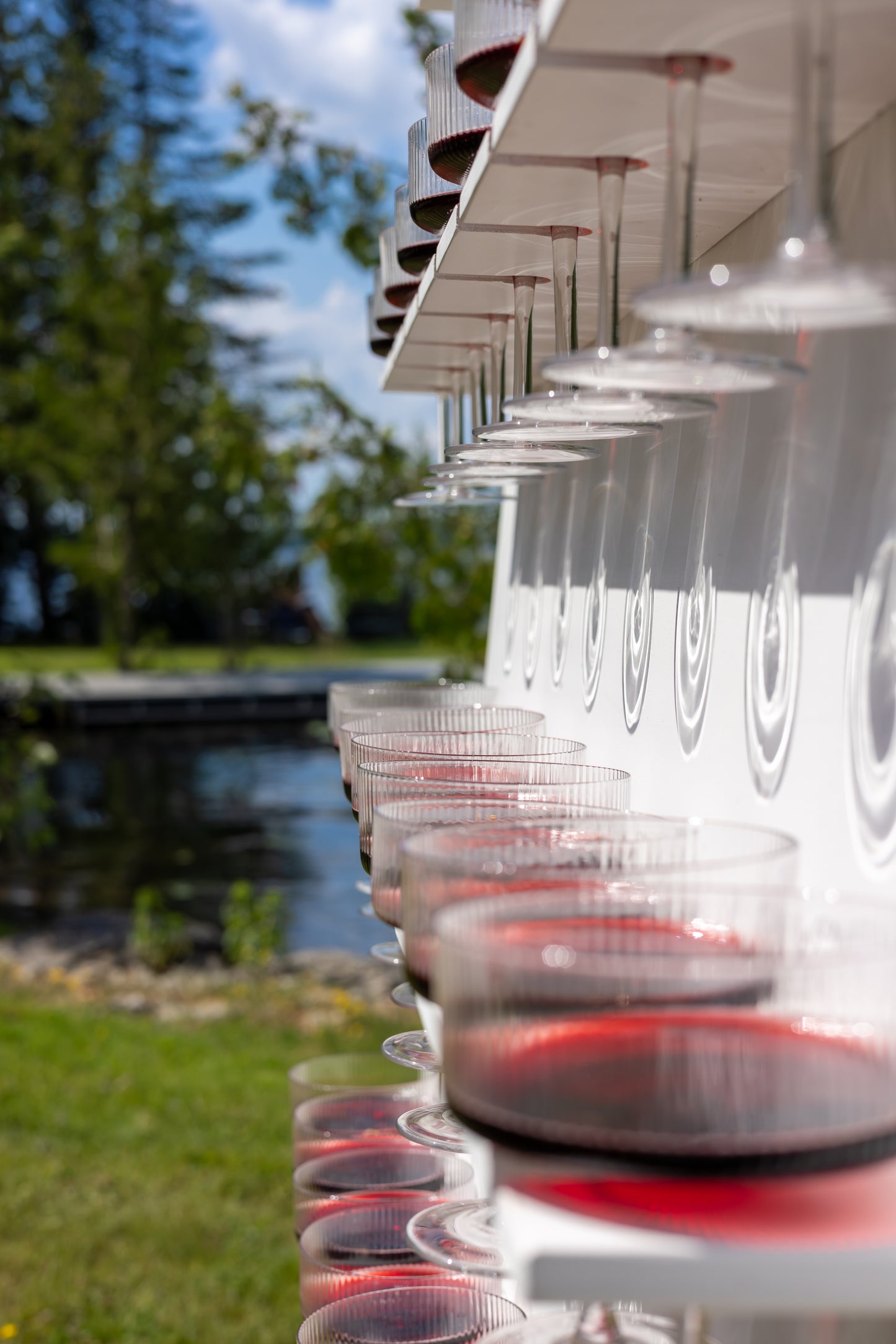 Row of clear plastic cups with red liquid on a table outdoors, with trees and sky in the background.