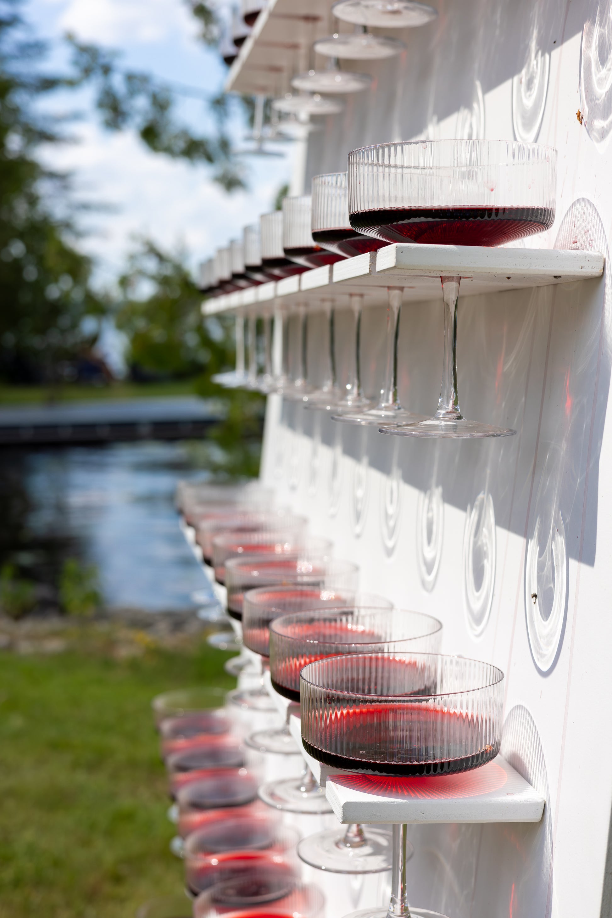 Row of wine glasses with red wine on a white outdoor shelf by a body of water.
