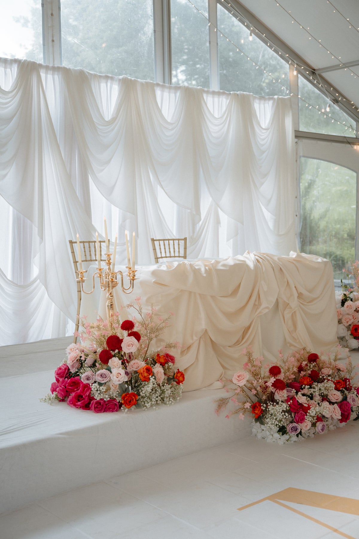 A styled bridal shower or wedding event space with draped white fabric, sheer curtains, gold chairs, and large floral arrangements in red, pink, and white on a raised platform.